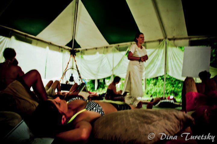 Ashley is walking in a crowded tent with people laying ont he floor. there is a buddha statue in the left corner of h the image. the tent is a green and white striped outdoor tent with white flowy fabric hanging down. Ashley is wearing an all white long dressMassage workshops in Philadelphia
