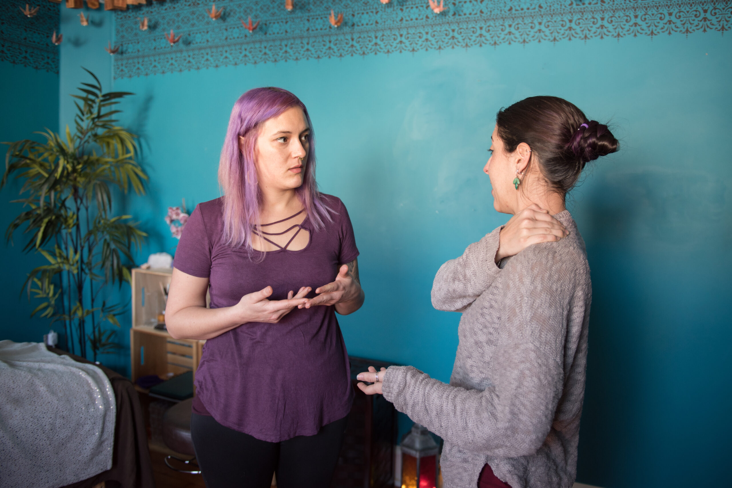 Two individuals in conversation, one with purple hair in a mauve top gesturing with hands, the other in a gray sweater holding their shoulder, in a room with a turquoise wall and a decorative border.