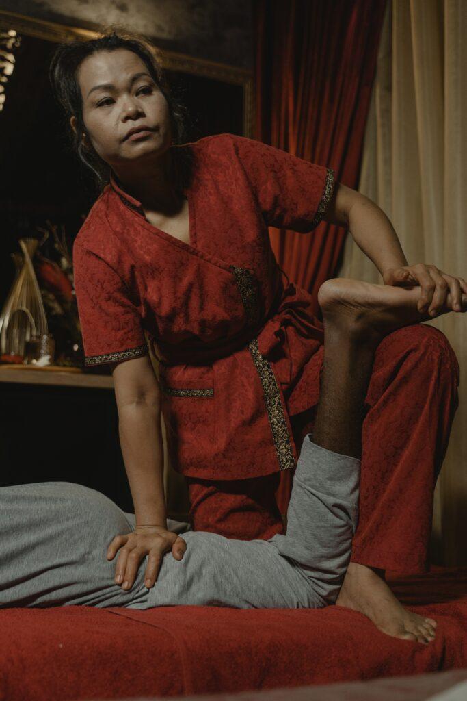 thai masage therapist is dressed in a red uniform witha. V-neck short sleeved top and red pants. there is a gold trim around the sleeves as well as the front pocket. She has her right hand on the hamstrings of a man that is face down. You can only see his left leg and glute. he is wearing grey sweatpants. The Thai massage therapist has his left leg bent at the knee as she pushed on his hamstring she is stabilizing the foot.