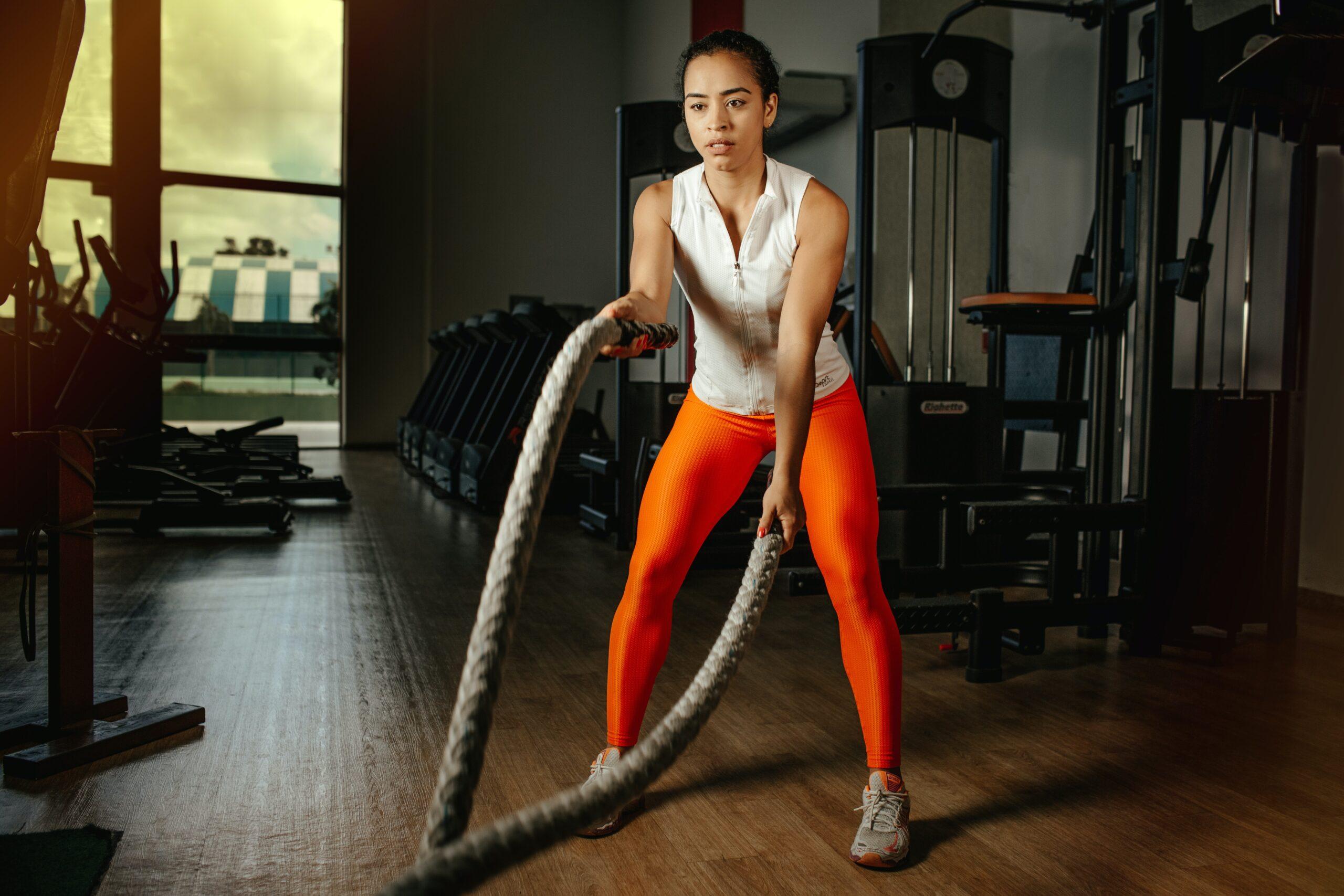 A focused woman in a white sleeveless top and orange leggings exercises with battle ropes in a gym, with gym equipment in the background.