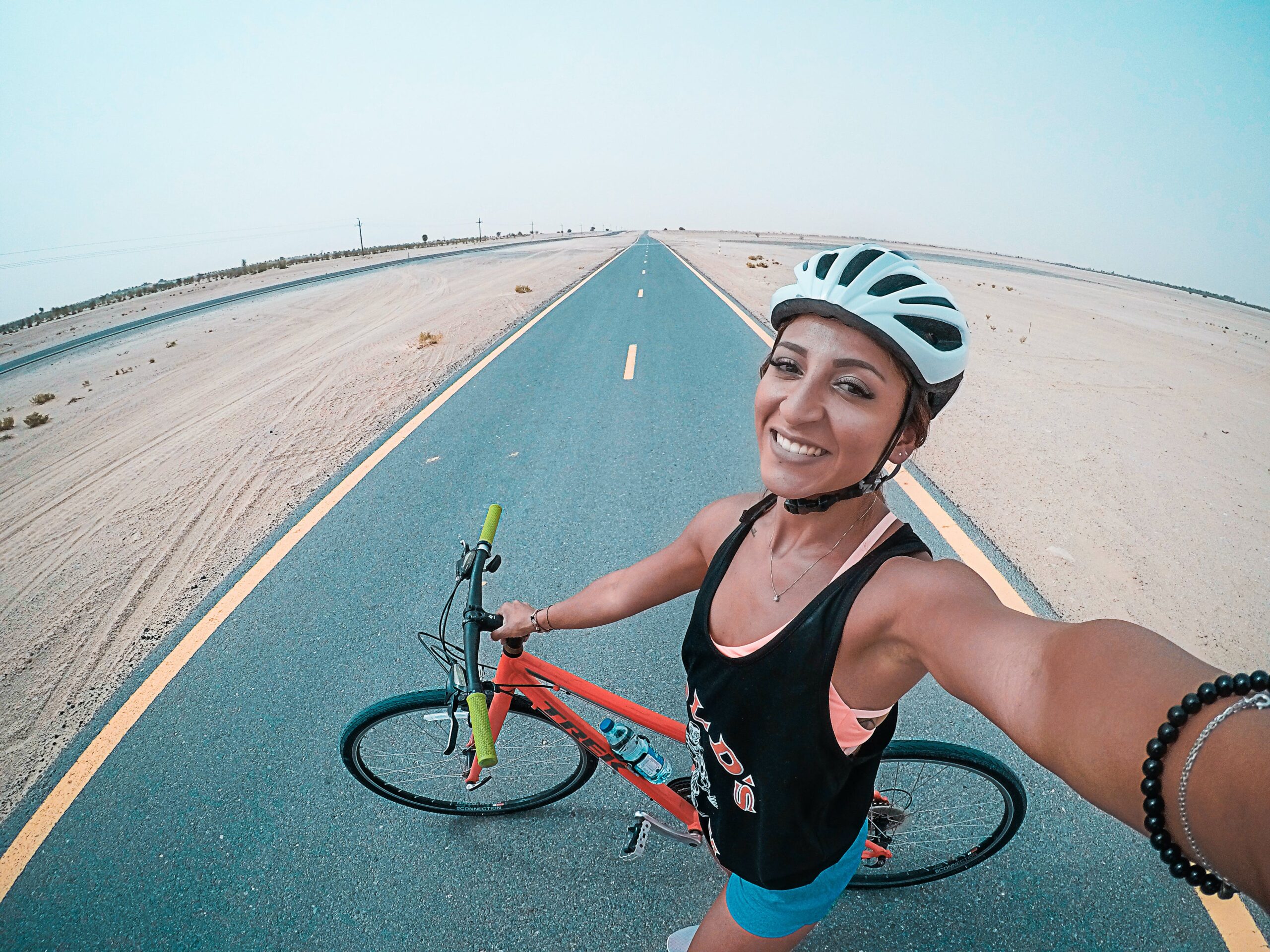 A happy cyclist takes a selfie on an empty desert road with her red bicycle, wearing a helmet, tank top, and shorts, with clear skies above.
